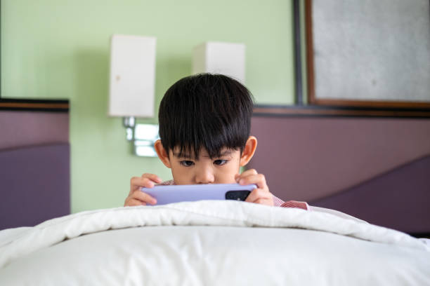 A little Asian boy lies on the bed playing with his phone.